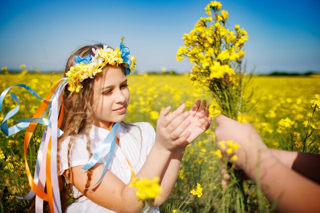 Mädchen im Kleid mit ukrainischem Kranz mit Blumen und Bändern nimmt aus den Händen eines unbekannten Jungen einen Strauß Rapsblumen