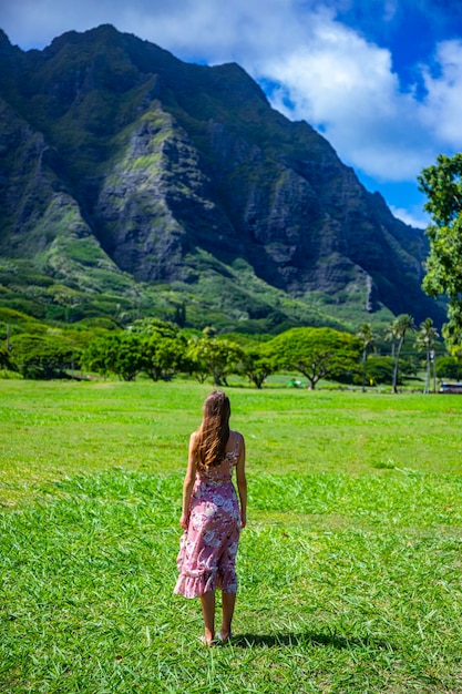 Mädchen im langen Kleid geht auf Gras am Kualoa Point auf Oahu, Hawaii, mit Blick auf den mächtigen grünen Berg