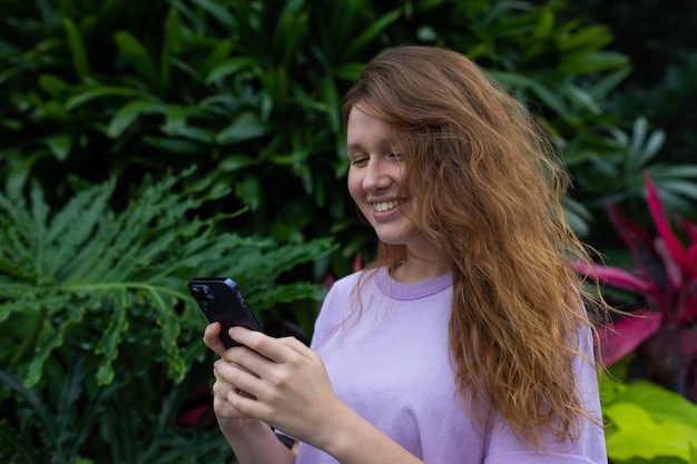 Mädchen im Park oder in der Natur nutzen ein Mobiltelefon, schauen auf den Bildschirm, lächeln und kommunizieren