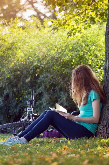 Mädchen im Park sitzt unter einem Baum und liest ein Buch. daneben steht ein Fahrrad und ein Rucksack