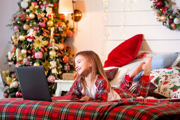 Mädchen im Pyjama liegt auf einem Bett auf rotem Plaid eines Weihnachtsbaumes und schaut auf den Monitor des Laptops.