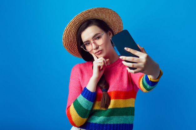 Mädchen im Regenbogenpullover hält den Finger auf die Wange und benutzt das Handy, um Selfies zu machen