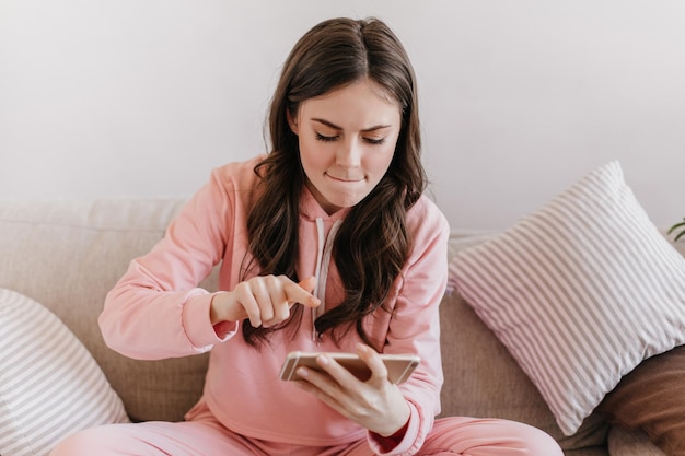 Mädchen im rosafarbenen Anzug mit Eifer drückt beim Spielen auf den Bildschirm des Telefons. Aufnahme einer Frau im Sport-Hoodie, die auf einem beigen Sofa sitzt, das von gestreiften Kissen umgeben ist