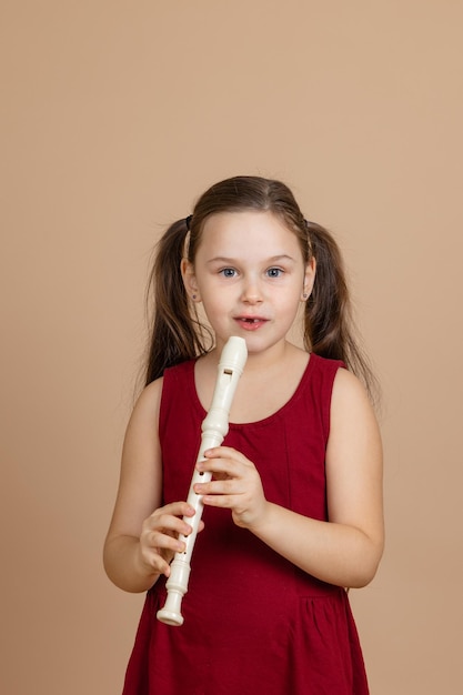 Mädchen im roten Kleid halten nachdenklich eine gemeinsame Flöte und kneifen Löcher auf dem beigen Hintergrund Lernen, Holzblasinstrumente zu spielen Flöte und Kinder ist ein Konzept der Entwicklung der Musikausbildung