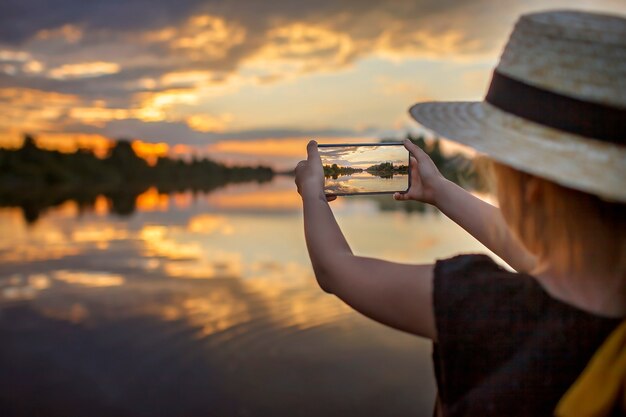 Mädchen im Sommerstrohhut, der bei Sonnenuntergang auf dem Seelebensstil lokale Reisen selfie für das Handy nimmt