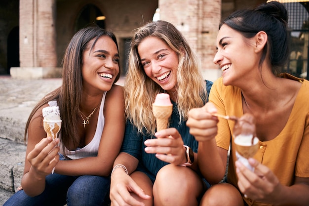 Mädchen im Teenageralter lachen und essen Eistüten, die draußen sitzen und ihren Sommerurlaub genießen