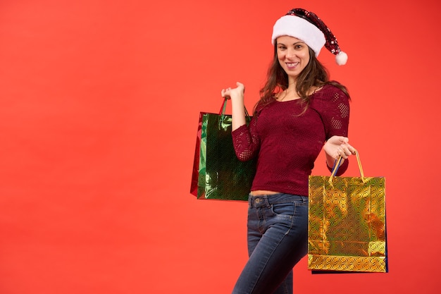 Mädchen im Weihnachtsmann-Hut, der mit Weihnachtseinkaufstaschen auf orange Hintergrund lächelt.