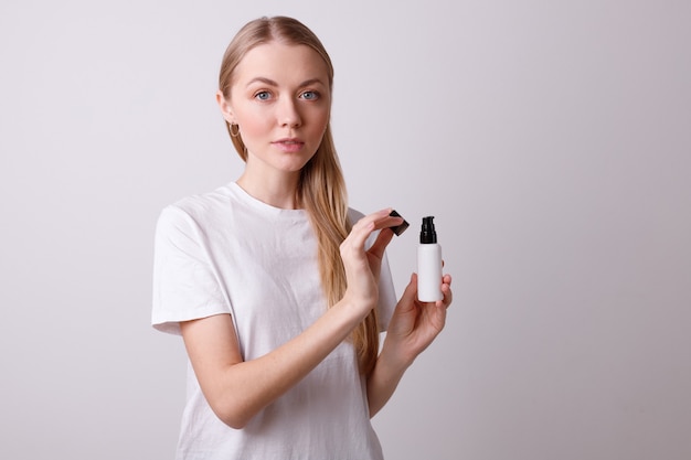 Mädchen im weißen T-Shirt hält eine Flasche Make-up an einer grauen Wand