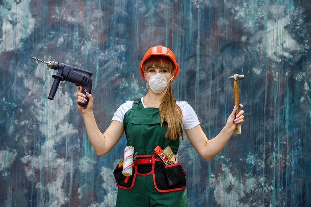 Mädchen in Baukleidung und Schutzausrüstung posiert mit Bohrer und Hammer auf grauer Wand.