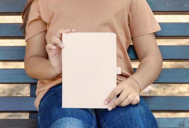 Foto mädchen in beige t-shirt sitzt auf einem bech und hält ein notizbuch in den händen