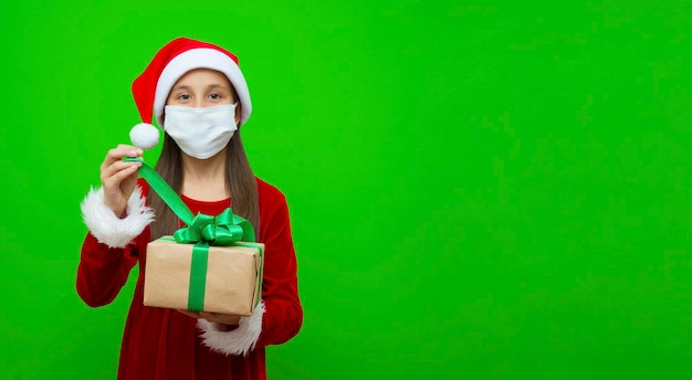 Mädchen in der medizinischen Maske des Sankt-Hutes in ihren Händen öffnet Geschenk für Weihnachtsgrünhintergrundfahne