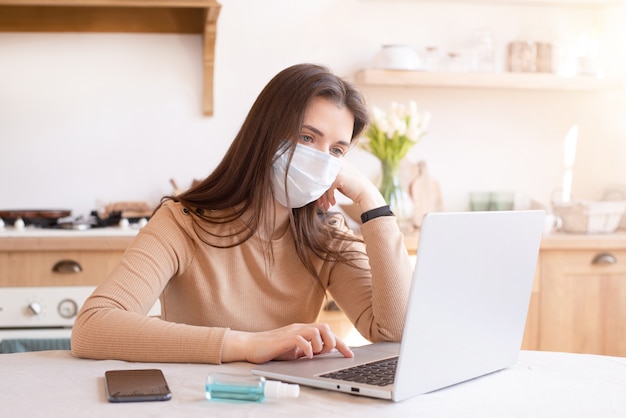 Mädchen in der medizinischen Maske, die am Laptop zu Hause in der Küche arbeitet. Coronavirus Isolation.