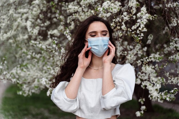 Mädchen in der medizinischen Maske nahe weißen blühenden Bäumen im Park. Outdoor-Wanderlandschaft in Quarantäne Coronavirus Covid-19-Zeitraum. Frühlingslebensstil.