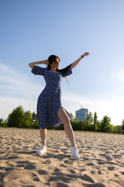 Mädchen in einem blauen Kleid, das auf dem Sand nahe dem Fluss aufwirft. Frau, die sonniges Wetter genießt.