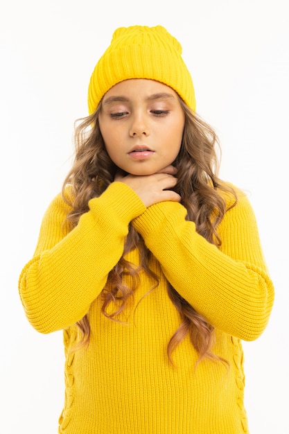 Mädchen in einem gelben Hut und einer Jacke hält Hände nahe der Kehle auf Weiß