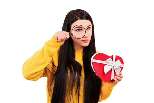 Mädchen in einem gelben Pullover mit einer Lupe in der Hand und einem Geschenk in Form eines Herzens, eine Konzeptsuchüberraschung isoliert auf weißem Hintergrund