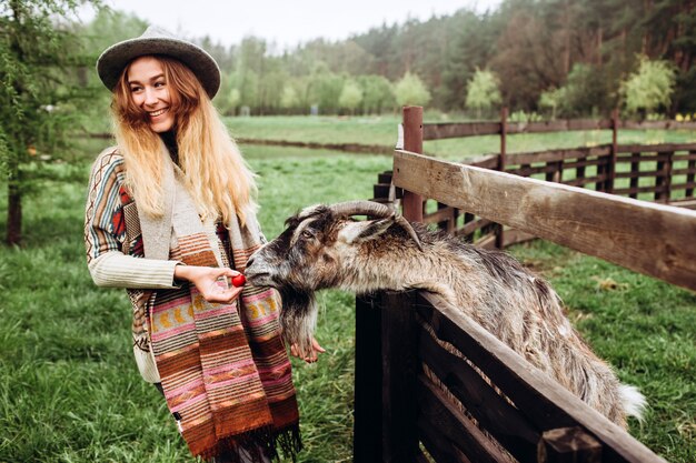 Mädchen in einem grauen Hut und in einem Pullover mit einem ethnischen Muster. Eine junge Frau füttert eine Ziege mit Gemüse in der Nähe eines Holzzauns auf einem Bauernhof. Ziege hautnah. Zoo Leben. Landwirtschaft
