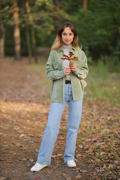 Mädchen in einem grünen Hemd und Blue Jeans im Herbstwald Mädchen, das ein Ahornblatt in ihren Händen hält