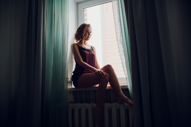 Mädchen in einem Hochzeitsschleier nahe einem Fenster in einem dunklen Raum