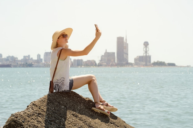 Mädchen in einem Hut macht ein selfie, das auf einem Felsen durch das Meer sitzt