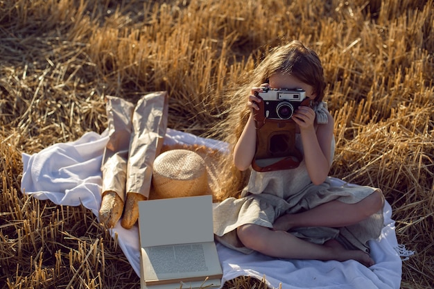 Mädchen in einem Kleid, das auf einem gemähten Feld mit einer Kamera auf einer Decke mit Brot und einem Buch sitzt