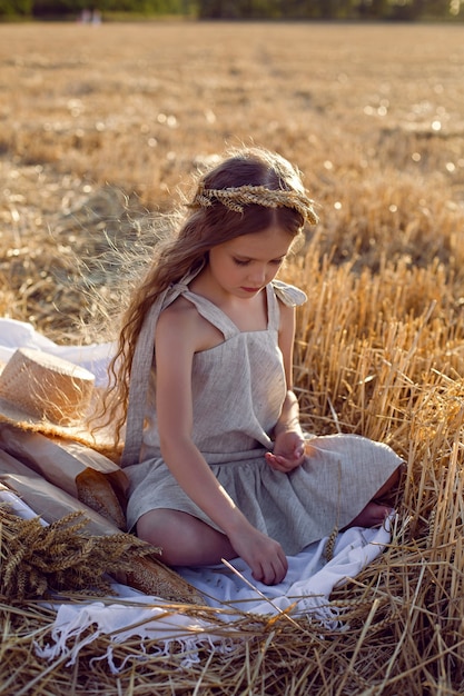 Mädchen in einem Kleid und einem Kranz auf ihrem Kopf sitzen auf einem gemähten Weizenfeld bei Sonnenuntergang im Sommer