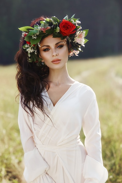 Mädchen in einem langen Kleid steht in einem Feld mit Kranz auf dem Kopf und Blumenstrauß in den Händen, schöne Frau in den Strahlen der Abendsonne im Herbst im Dorf