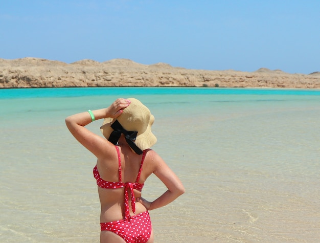 Mädchen in einem roten Badeanzug und einem Strandhut steht der Sonne im Meer gegenüber. Sonnenbäder stehen im blauen transparenten Wasser.