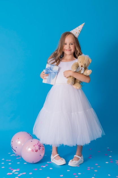 Mädchen in einem schönen, flauschigen weißen Kleid hält einen Teddybären und eine Geschenkbox in ihren Händen