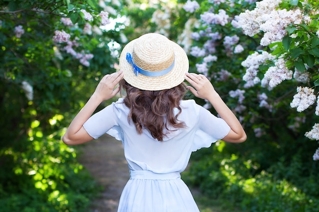 Mädchen in einem Strohhut mit blauem Band an einem Frühlingsnachmittag. Rückansicht. Trendy lässiges Sommer- oder Frühlingsoutfit. Frau im Strohhut eines Bootsmanns. Konzept der weiblichen Frühlingsmode. Blühende Fliederbüsche