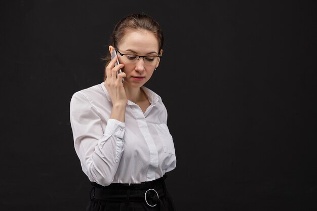 Mädchen in einem weißen Hemd in der Brille spricht auf einem Smartphone auf einem schwarzen Hintergrund.