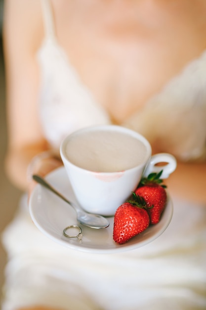 Mädchen in einem weißen Kleid hält in ihren Händen eine Untertasse mit einer Tasse Cappuccino zwei Erdbeeren a