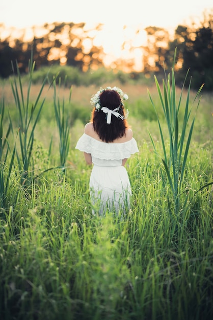 Mädchen in einem weißen Kleid mit einem Blumenkranz steht zurück auf dem Gebiet im Sommer