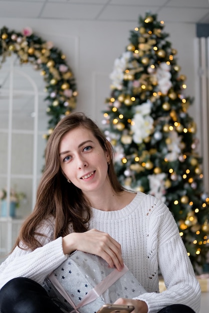 Mädchen in einem weißen Pullover sitzt mit einem Weihnachtsgeschenk auf dem Hintergrund eines Weihnachtsbaums.