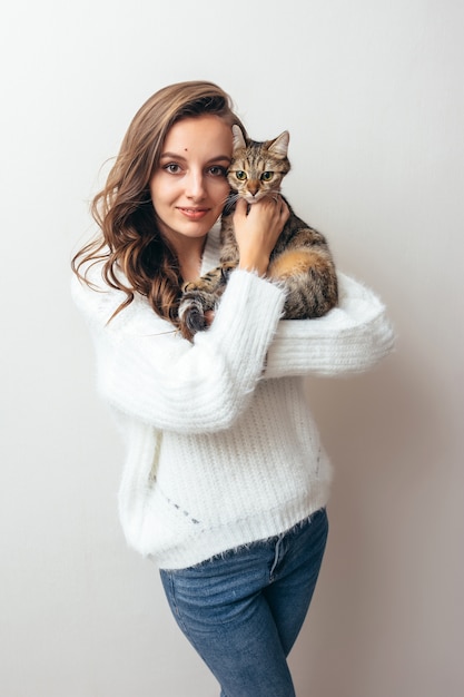 Mädchen in einem weißen Pullover umarmt eine graue Katze auf einem weißen