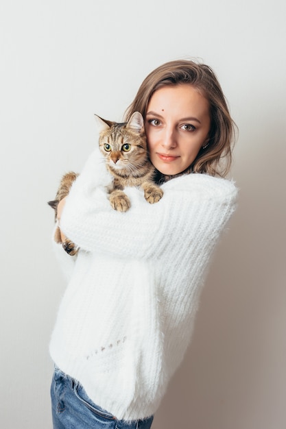 Mädchen in einem weißen Pullover umarmt eine graue Katze auf einem weißen