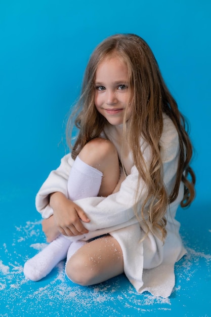 Mädchen in einem weißen Pullover und Hut bläst künstlichen Schnee von der Palme im Studio auf blauem Hintergrund