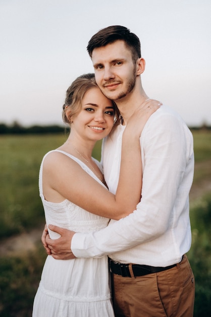 Mädchen in einem weißen Sommerkleid und ein Mann in einem weißen Hemd auf einem Spaziergang bei Sonnenuntergang mit einem Blumenstrauß in einem Dorf außerhalb der Stadt