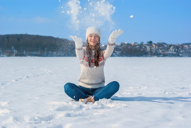 Mädchen in einem weißen Winterpullover sitzt mit gekreuzten Beinen im Schnee wirft den Schnee auf