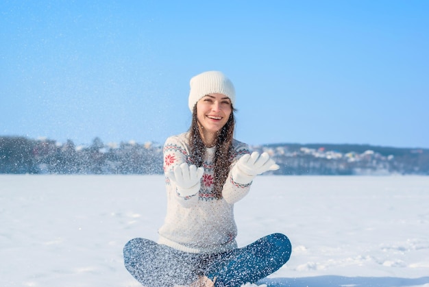 Mädchen in einem weißen Winterpullover sitzt mit gekreuzten Beinen im Schnee wirft den Schnee auf