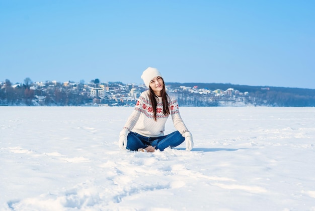 Mädchen in einem weißen Winterpullover sitzt mit gekreuzten Beinen im Schnee wirft den Schnee auf