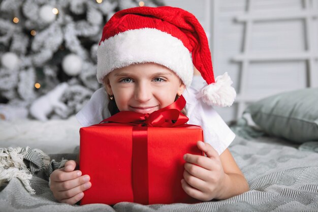 Mädchen in einer roten Mütze mit einem Weihnachtsgeschenk. Sie liegt auf dem Bett, umarmt die Schachtel vor dem Hintergrund des Weihnachtsbaums.
