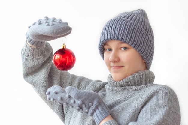 Mädchen in einer Weihnachtsmütze, grauem Pullover und Handschuhen hält eine rote Ballondekoration für den Weihnachtsbaum