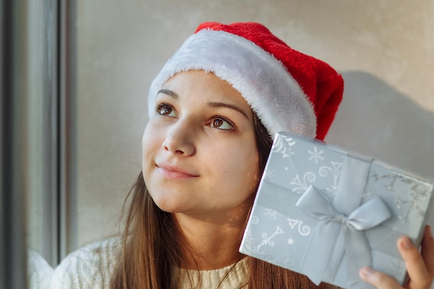 Foto mädchen in einer weihnachtsmütze mit einer geschenkbox am fenster nahaufnahme