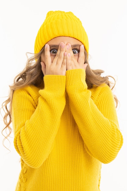 Foto mädchen in gelbem hut und pullover bedeckte ihr gesicht mit den händen und schaut durch die schlitze an der weißen wand