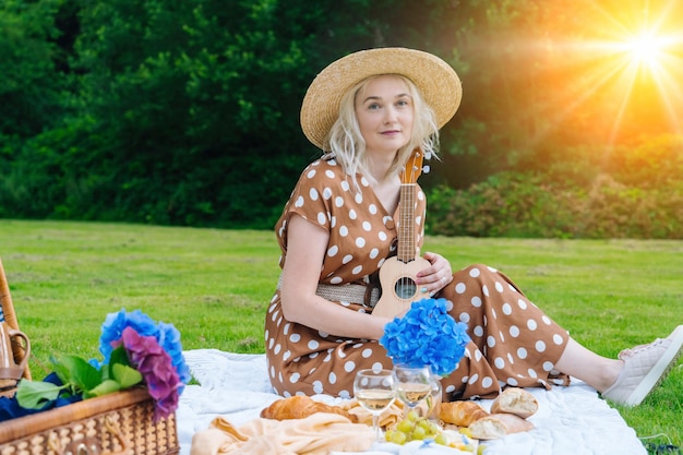 Mädchen in gepunktetem Kleid und Hut, das auf einer weißen Strickpicknickdecke sitzt, spielt Ukulele und trinkt Wein