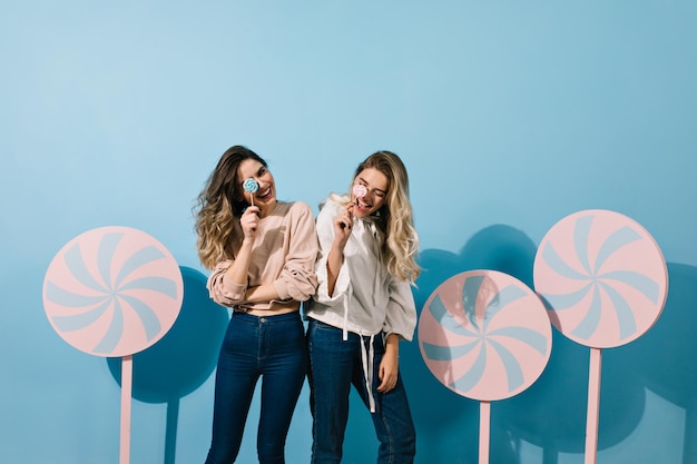 Mädchen in Jeans posieren mit Süßigkeiten Studioaufnahme von Freunden, die mit Lutschern auf blauem Hintergrund stehen