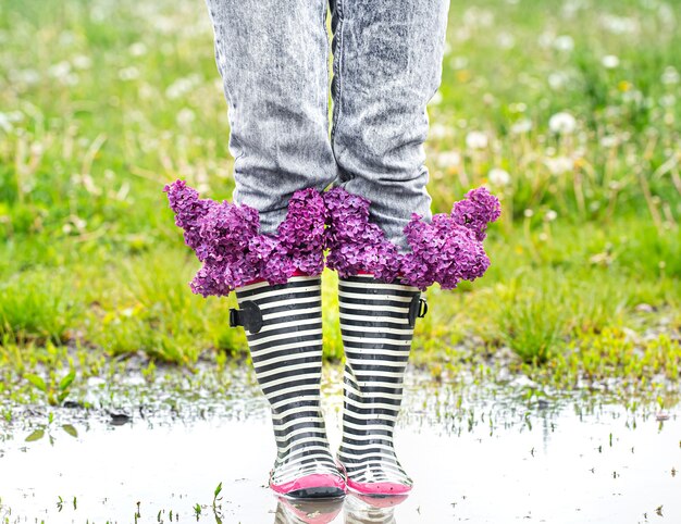 Mädchen in Jeans und gestreiften Gummistiefeln mit einem Strauß frischer lila Blumen in einer Pfütze unter dem Gras.