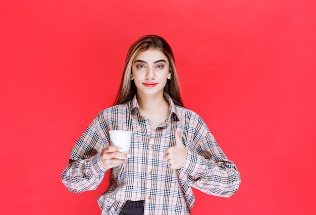 Mädchen in kariertem Hemd, das eine weiße Einweg-Kaffeetasse hält und den Geschmack genießt