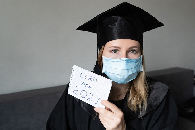 Mädchen in medizinischer Maske mit Abschlusshut, der eine Klasse von einundzwanzig Notizbüchern hält